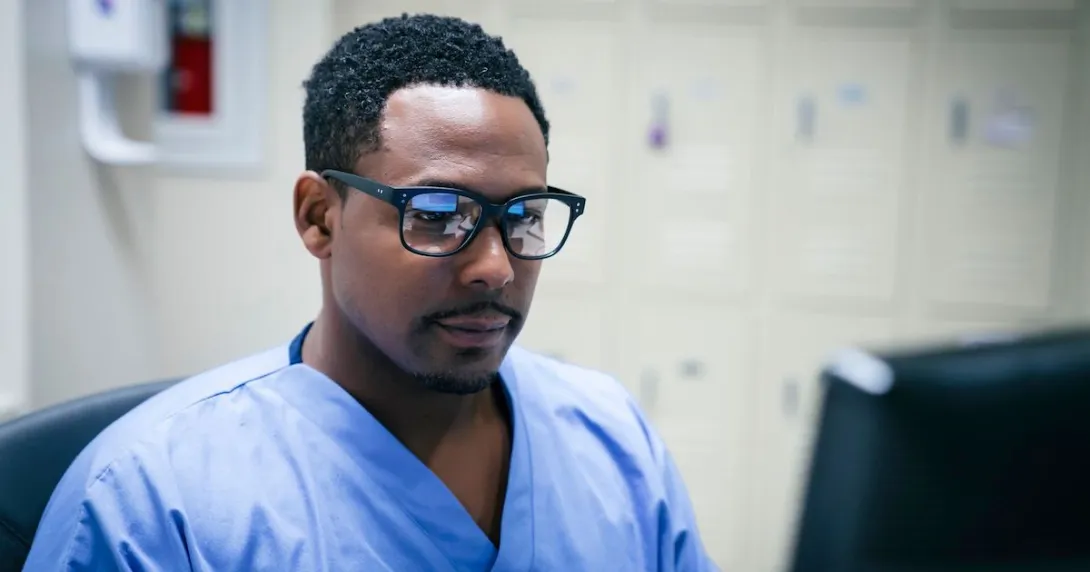 Healthcare provider wearing glasses with blue scrubs on sitting at a computer Healthcare provider wearing glasses with blue scrubs on sitting at a computer