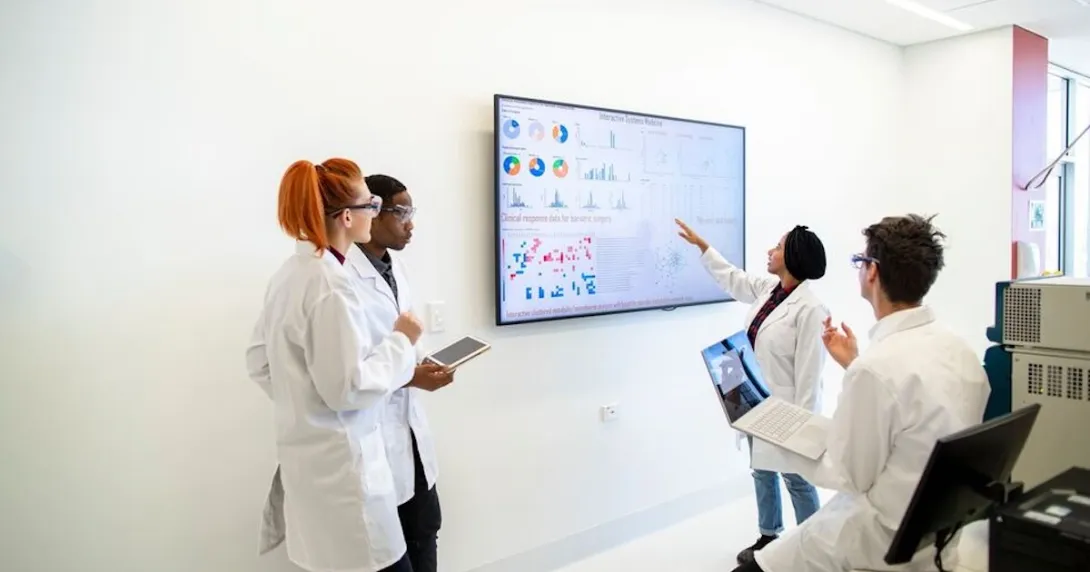 Four healthcare providers in a room looking at a screen with data on it Four healthcare providers in a room looking at a screen with data on it
