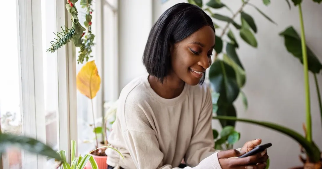 A person using a smartphone sitting by a window and surrounded by plants A person using a smartphone sitting by a window and surrounded by plants